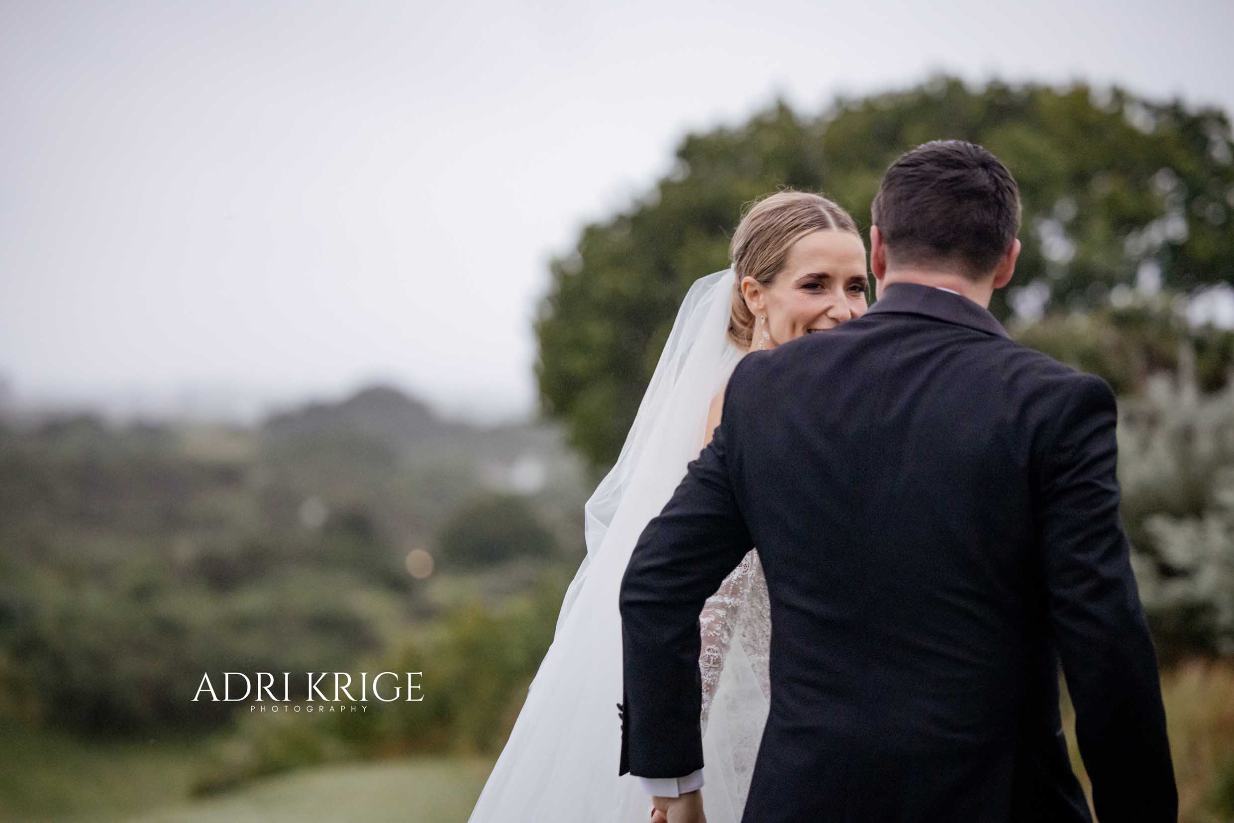 Smiling bride in a long veil looking over her groom’s shoulder as they walk hand in hand through misty green hills, wedding photography by Adri Krige, Eastern Cape wedding photographer