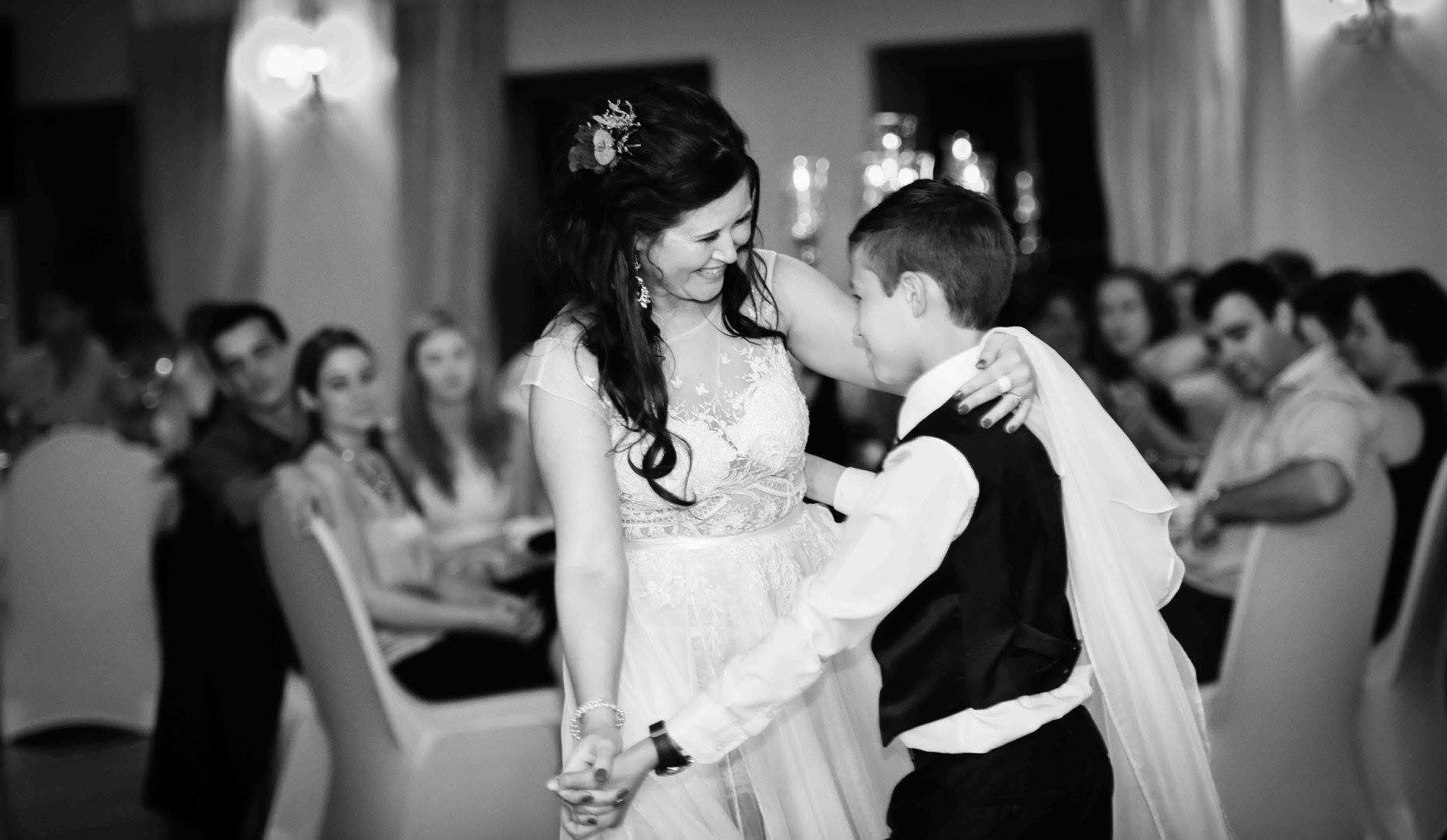 Black and white photo of a laughing bride with flowers in her hair dancing with a young boy in a waistcoat at the wedding reception as guests look on, wedding photography by Adri Krige, Eastern Cape wedding photographer