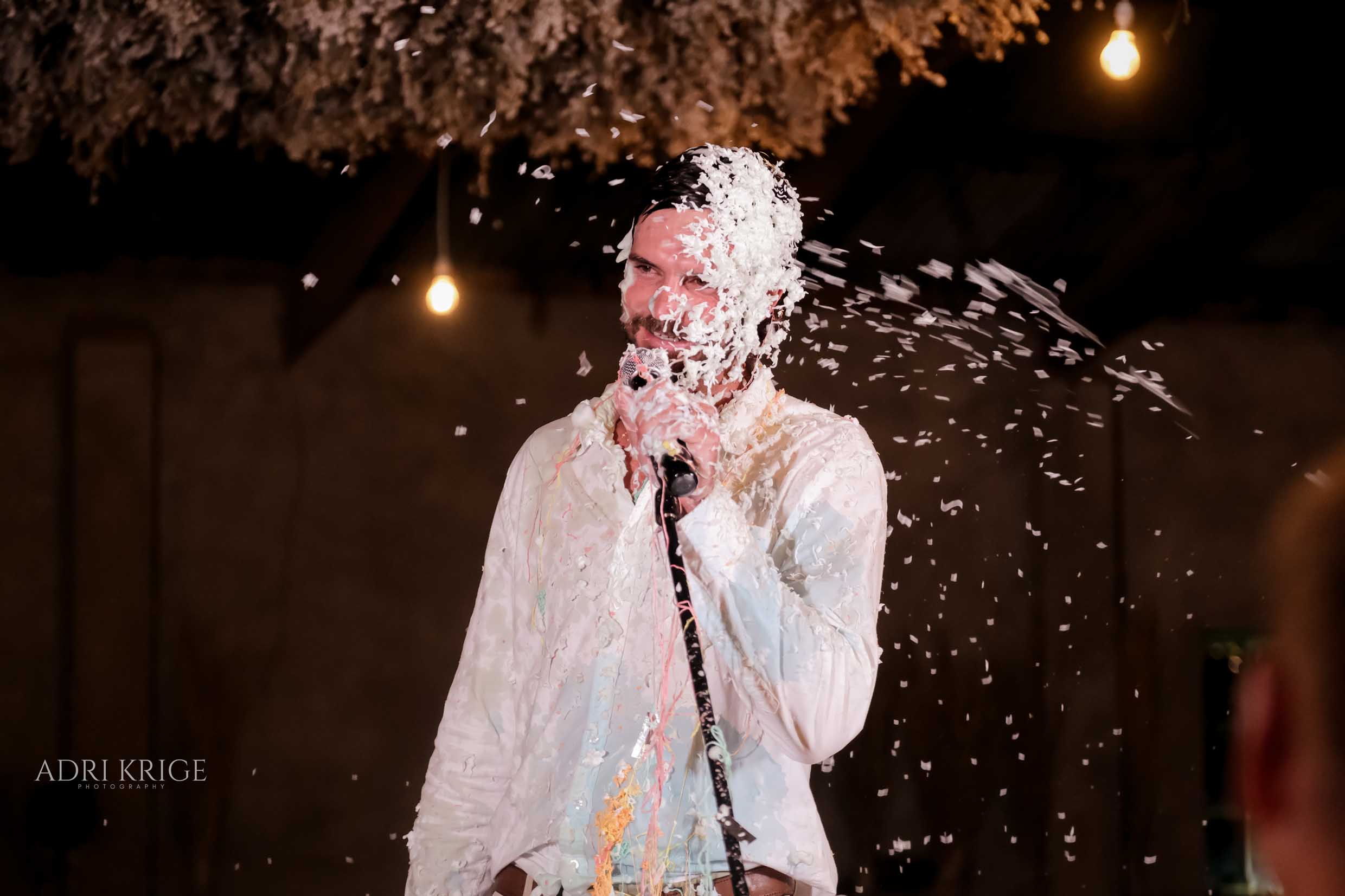 Smiling man in a white shirt holding a microphone at the wedding reception, laughing as guests cover him in party string spray under warm hanging lights, wedding photography by Adri Krige, Eastern Cape wedding photographer