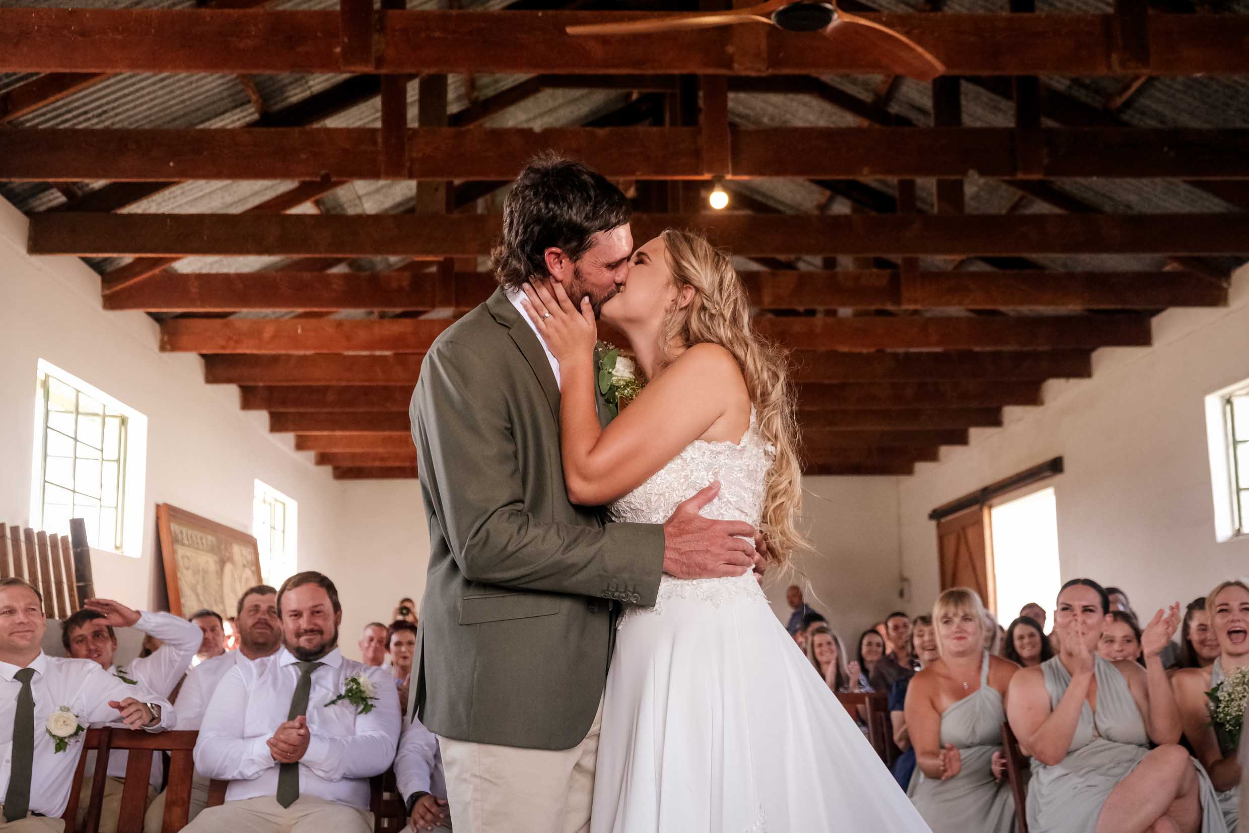 Bride and groom sharing their first kiss as husband and wife in a rustic farm barn with timber beams, as cheering guests, groomsmen and bridesmaids clap, wedding photography by Adri Krige, Eastern Cape wedding photographer