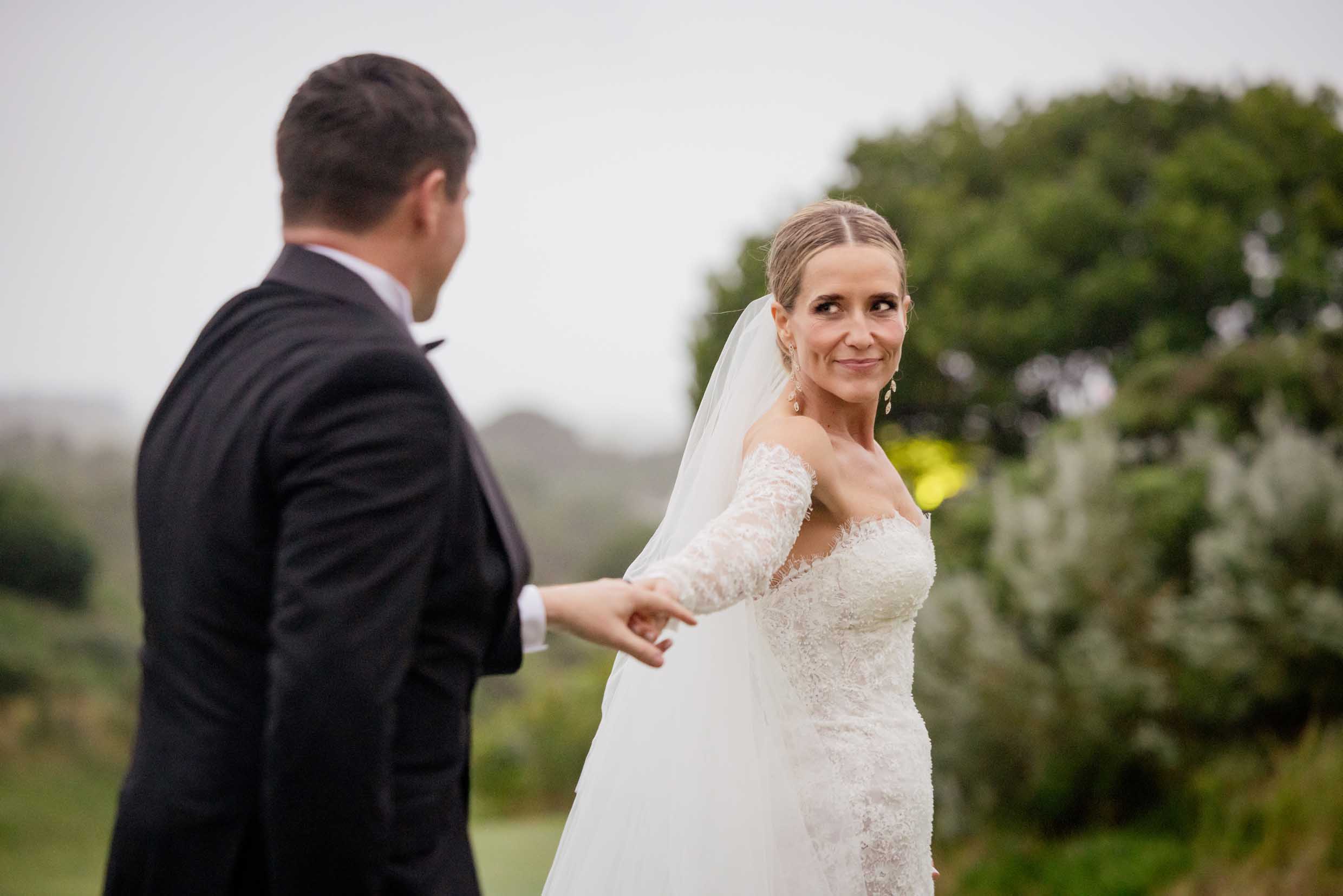 Smiling bride in an off-the-shoulder lace gown and long veil glancing back as she leads her groom in a black tuxedo by the hand through green gardens, wedding photography by Adri Krige, Eastern Cape wedding photographer