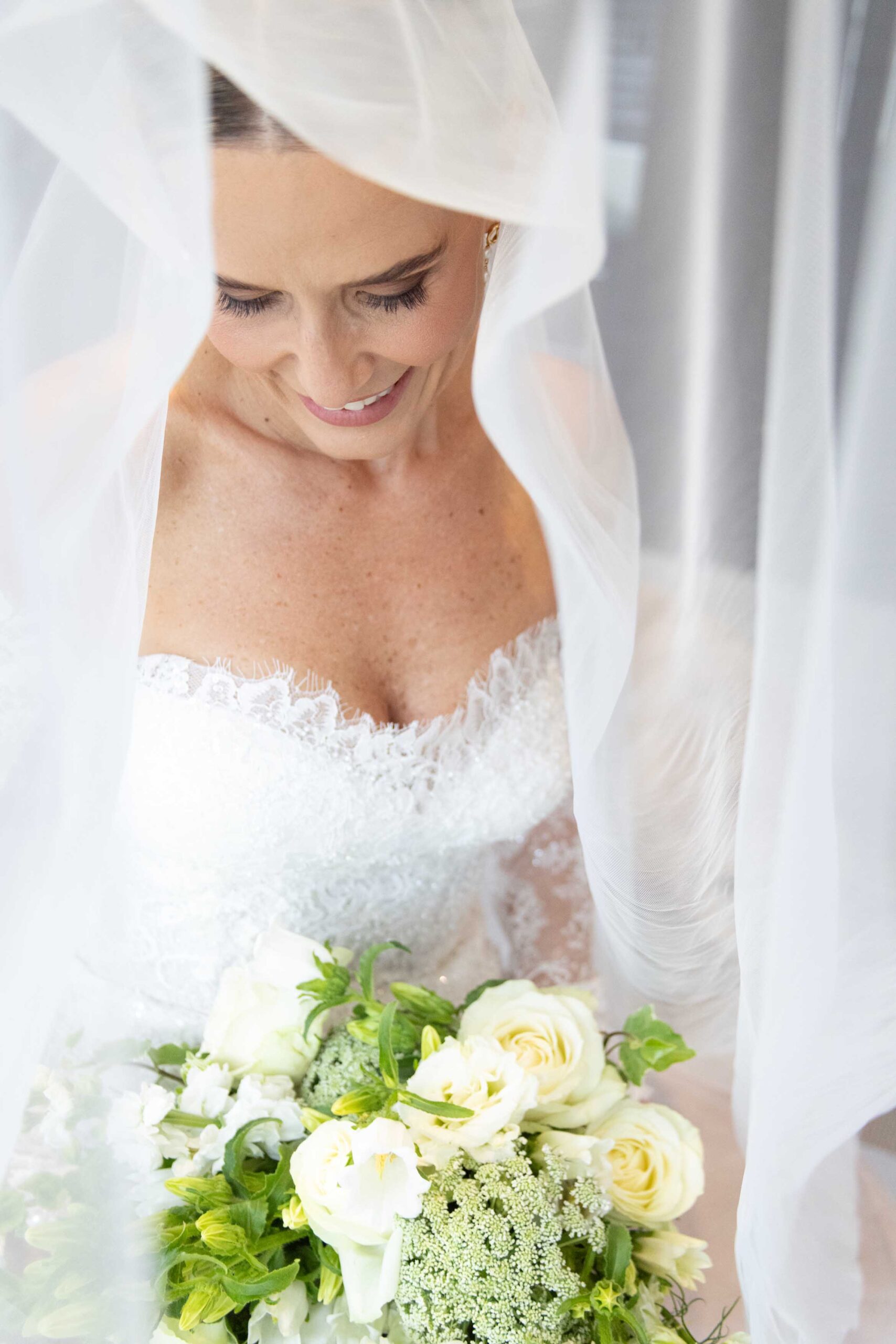 Smiling bride looking down beneath her sheer veil in an off-the-shoulder beaded lace gown, holding a bouquet of white roses and green blooms, bridal photoshoot by Adri Krige, Eastern Cape wedding photographer