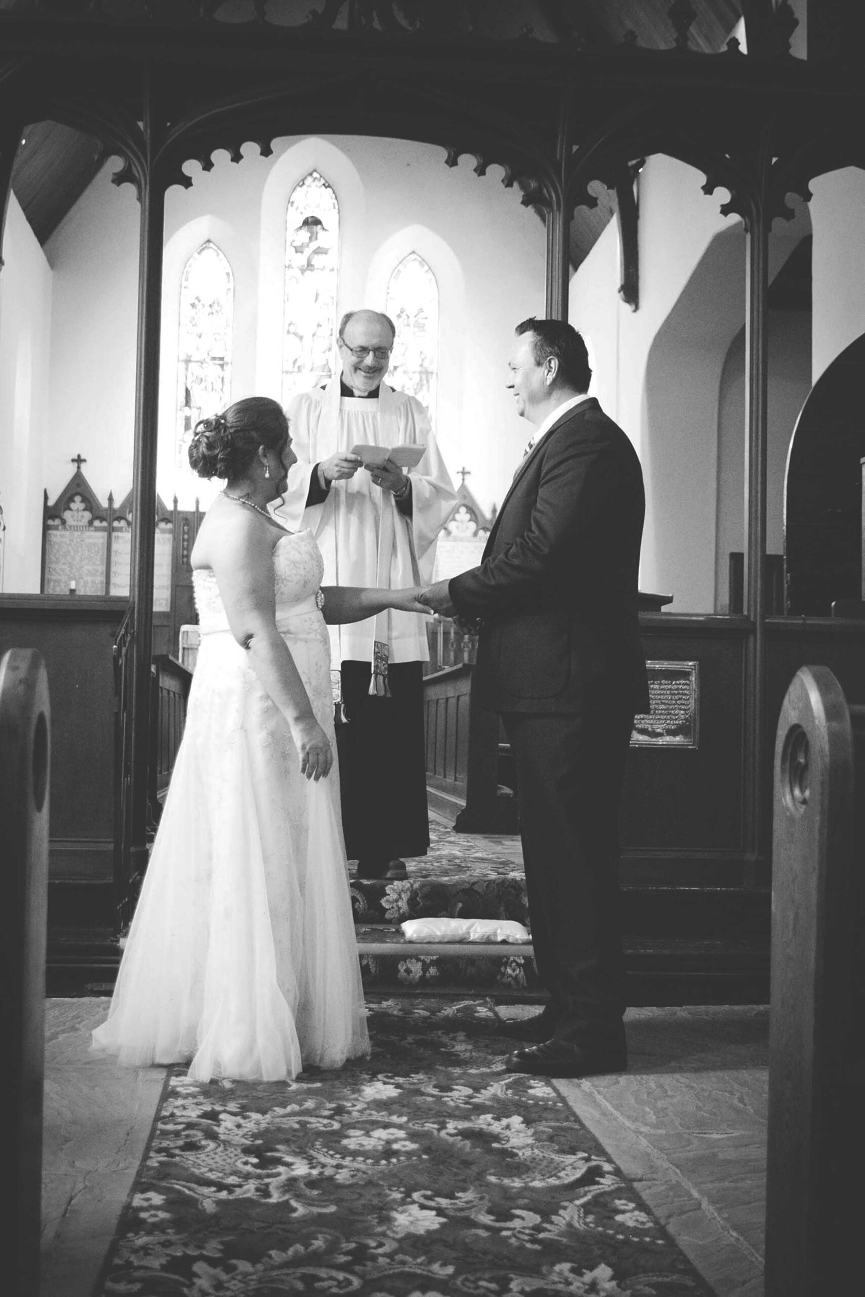 Black and white photo of a bride and groom holding hands at the altar of a small church as the smiling minister reads their vows, small wedding photography by Adri Krige, Eastern Cape wedding photographer