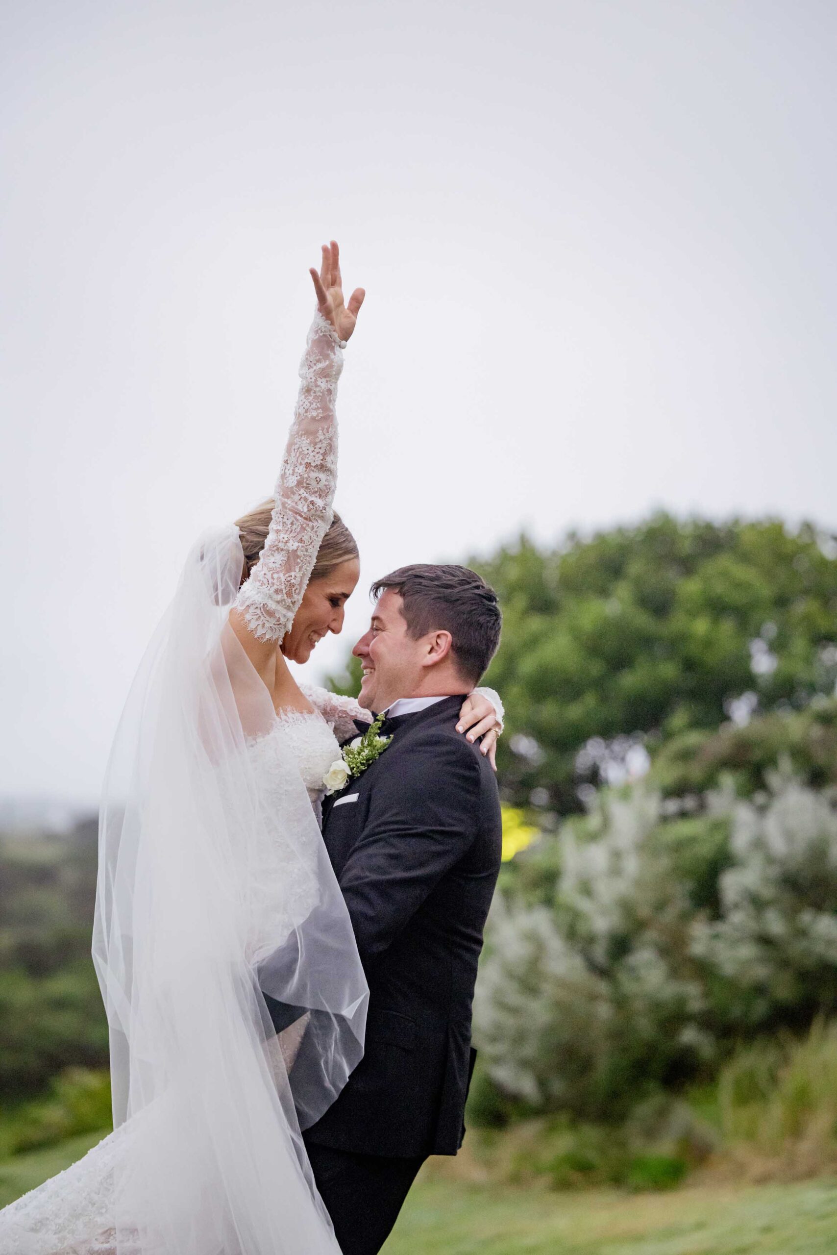Bride and groom celebrating, arm raised in joy Joyful bride in a long-sleeved lace gown and veil raising one arm in celebration as her laughing groom in a black tuxedo lifts her, with green coastal bush behind them, photographed by Adri Krige, wedding photographer St Francis Bay