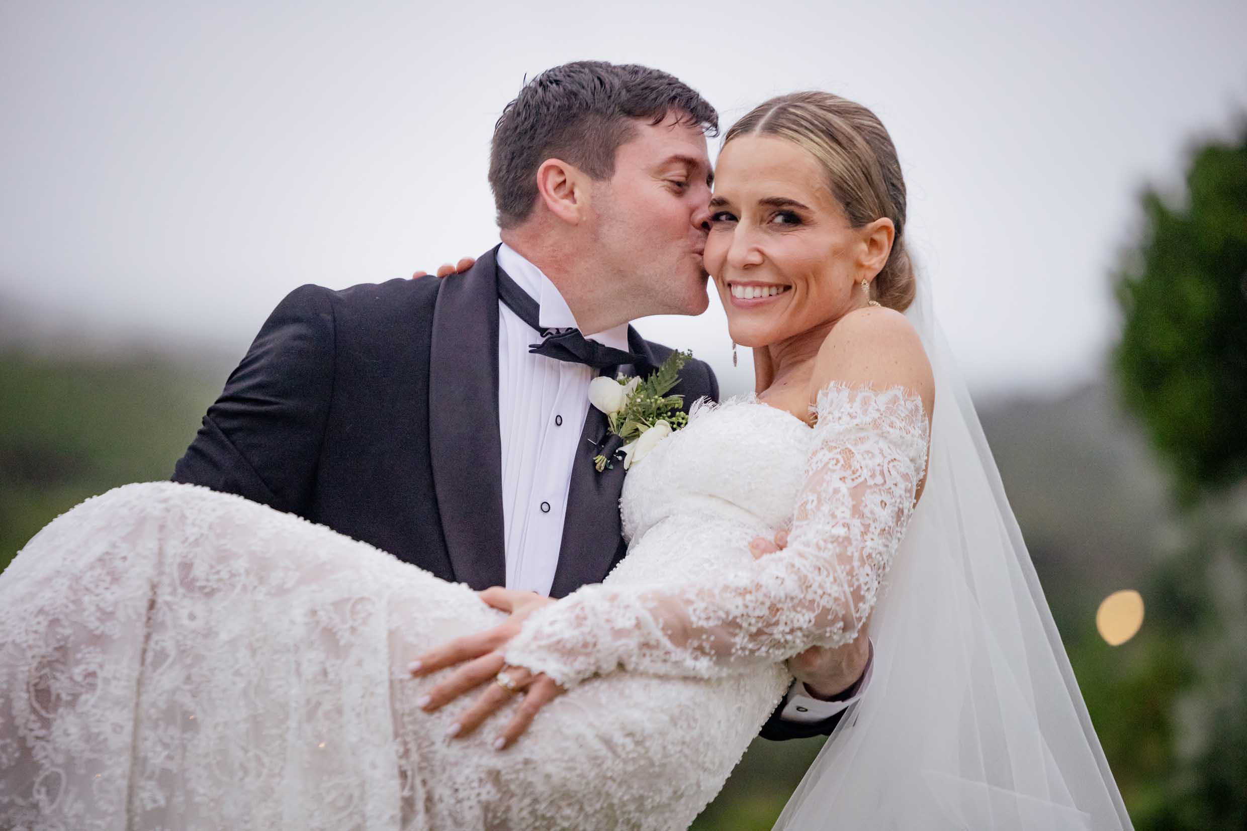 St Francis Links wedding, groom carrying his smiling bride Groom in a black tuxedo kissing the cheek of his smiling bride as he carries her in her off-the-shoulder lace gown and veil, on their wedding day at St Francis Links, photographed by Adri Krige, wedding photographer St Francis Bay