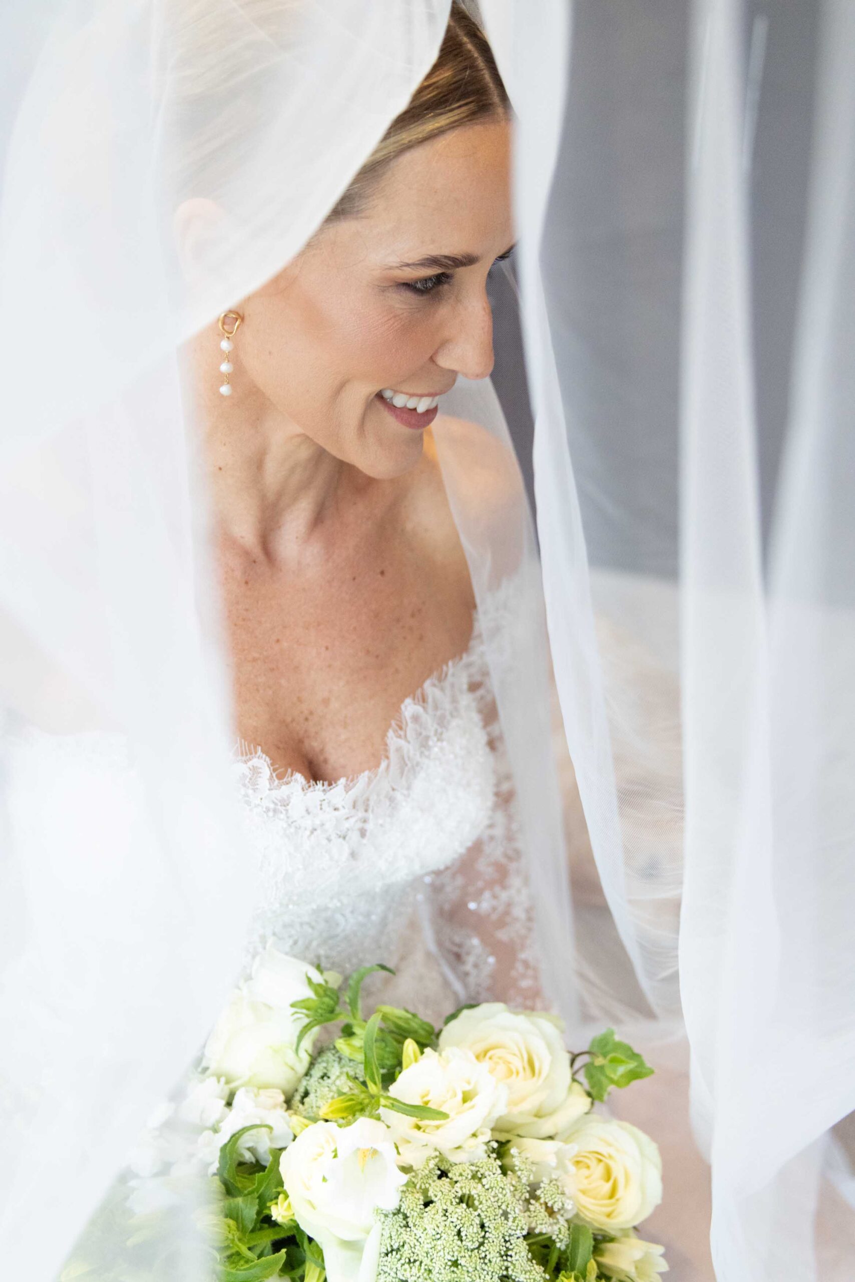 St Francis Links wedding, bride smiling beneath her veil Smiling bride with pearl drop earrings looking down beneath her sheer veil, holding a bouquet of white roses and green blooms, on her wedding day at St Francis Links, bridal photography by Adri Krige, St Francis Bay wedding photographer