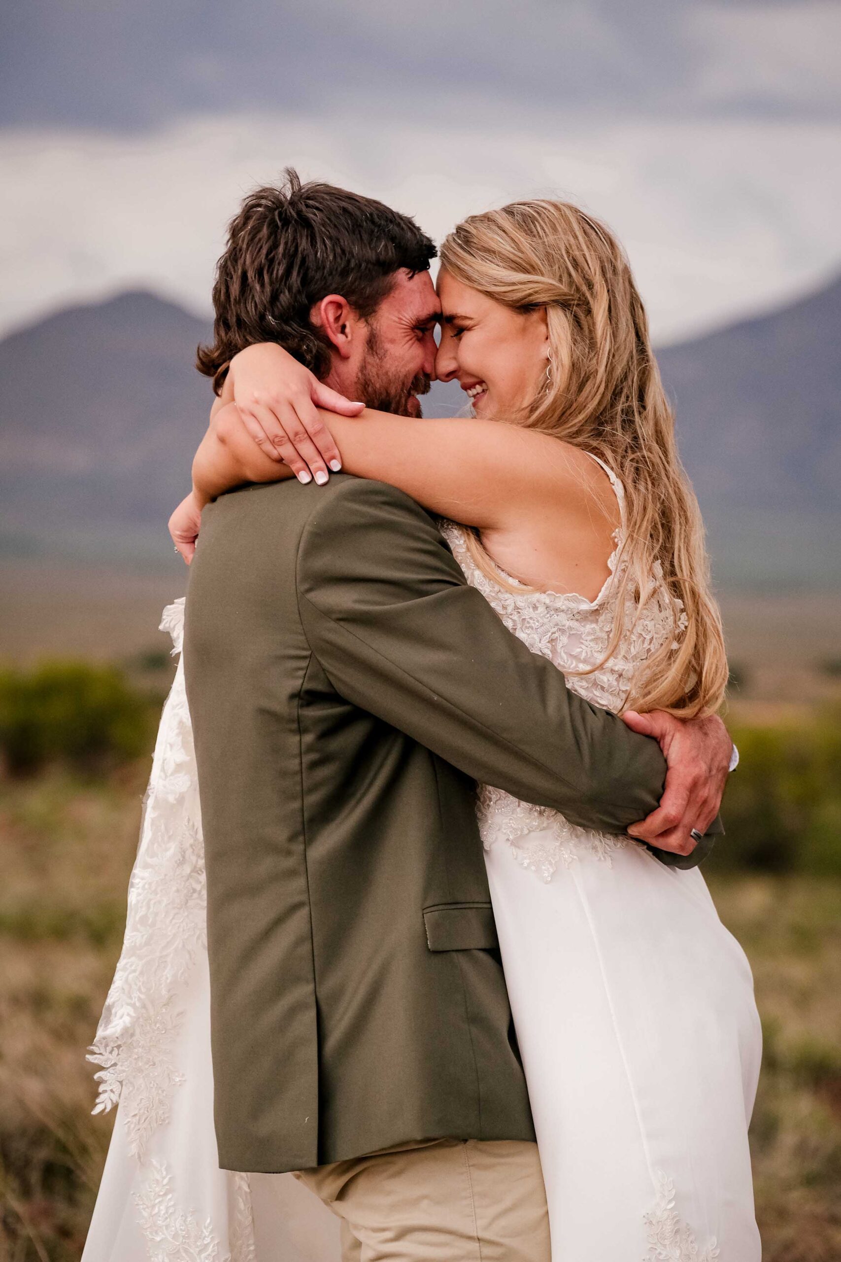 Laughing bride and groom in a tight embrace, foreheads together, she in a lace wedding gown and he in an olive green jacket, with mountains and open veld behind them, intimate wedding photography by Adri Krige, Eastern Cape wedding photographer