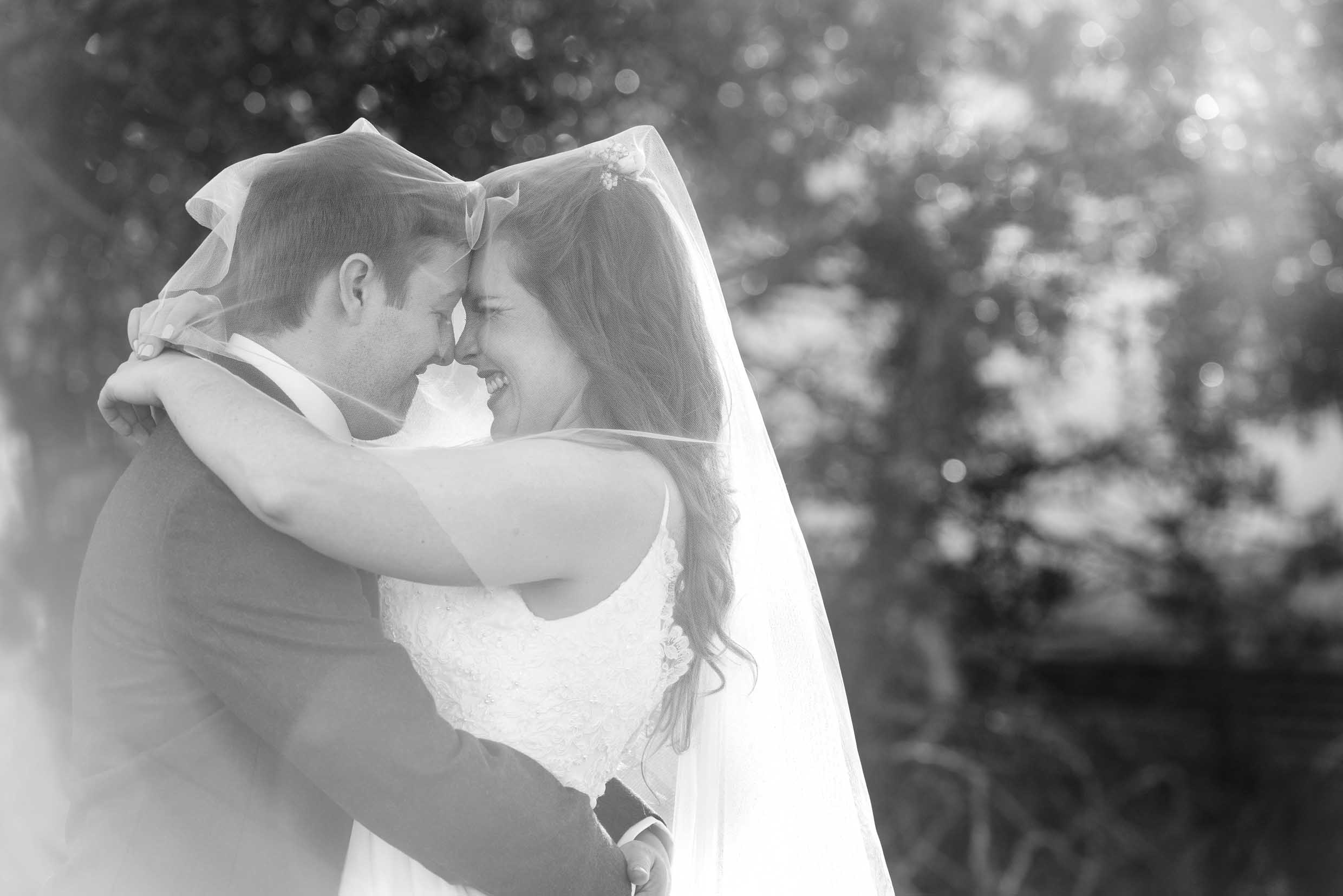 Black and white photo of a laughing bride and groom wrapped together under her veil, foreheads touching in soft backlight, photographed by Adri Krige, Eastern Cape wedding photographer