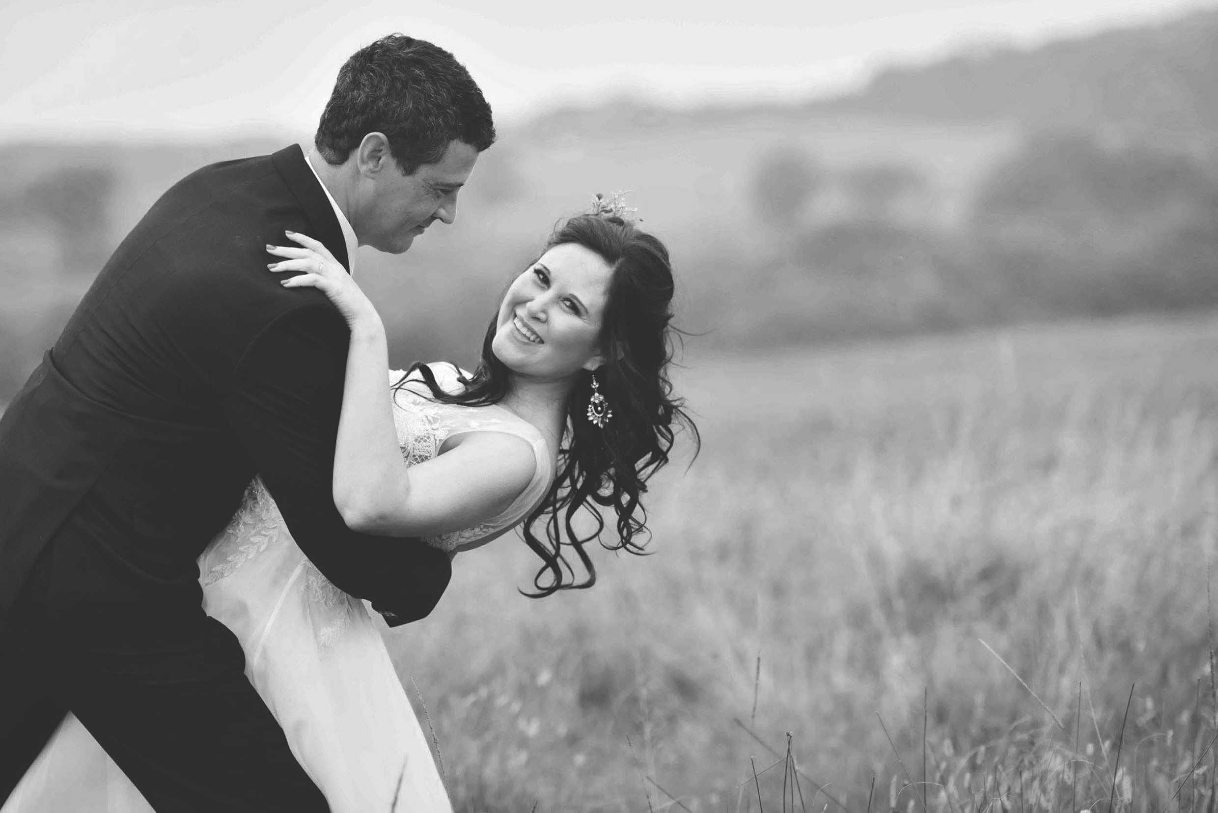 Black and white photo of a groom in a dark suit dipping his smiling bride in a lace wedding gown in a field of tall grass, wedding photography by Adri Krige, Eastern Cape wedding photographer