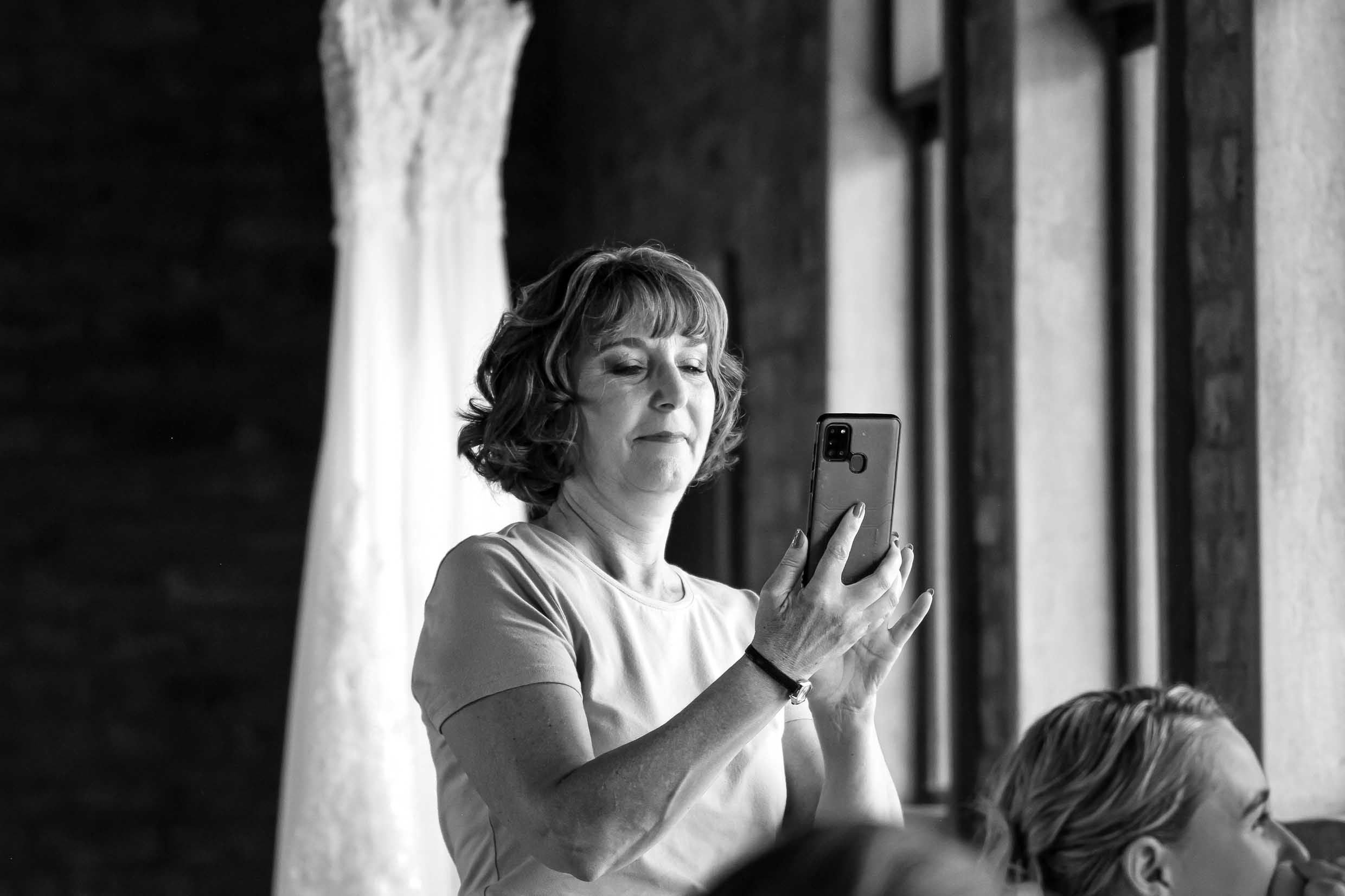 Black and white photo of a smiling woman taking a photo on her phone while the bride gets ready, with the wedding dress hanging in the background, wedding photography by Adri Krige, Eastern Cape wedding photographer