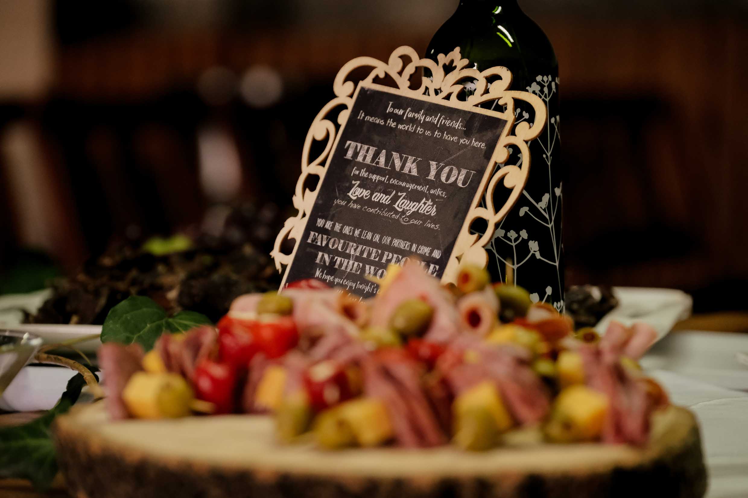 Chalkboard thank-you sign in a carved wooden frame beside a wine bottle on a wedding reception table, with a platter of cold meats, cheese and olives in front, wedding detail photography by Adri Krige, Eastern Cape wedding photographer