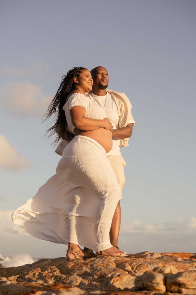 Expectant couple standing together on orange lichen-covered coastal rocks at sunset, her white skirt and long braids blowing in the wind as he holds her from behind with their hands on her bare bump, maternity photoshoot by Adri Krige Photography