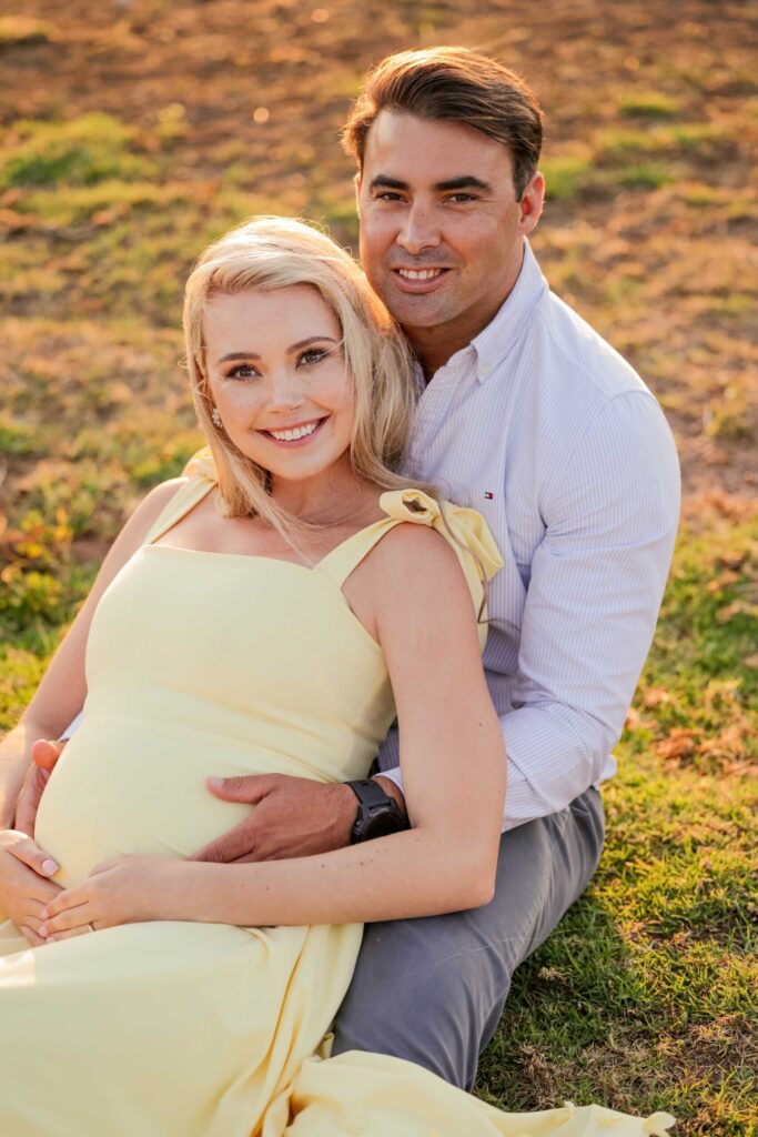 Smiling expectant couple sitting together on the grass, the mother in a pale yellow dress leaning back against her partner as they both rest their hands on her bump in warm evening light, maternity photoshoot by Adri Krige Photography