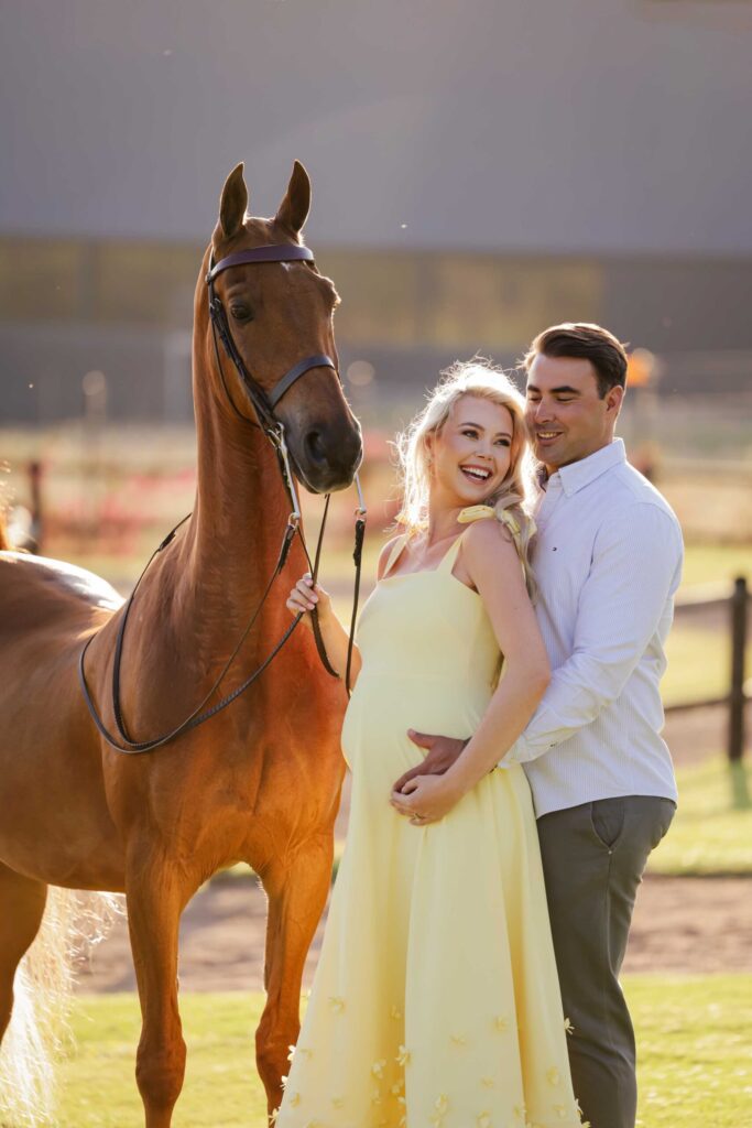 Laughing expectant mother in a yellow dress held from behind by her smiling partner, both hands resting on her bump, as she holds the reins of their chestnut horse beside a paddock fence at golden hour, maternity photoshoot by Adri Krige Photography
