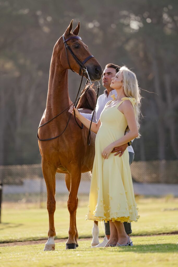 Expectant mother in a pale yellow dress with flower appliqués and her partner standing beside a tall chestnut horse in a bridle, both gazing up at it as he wraps an arm around her bump, maternity photoshoot by Adri Krige Photography