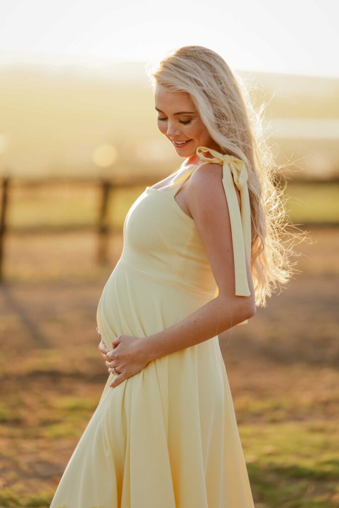Smiling expectant mother with long blonde hair in a flowing pale yellow dress with tied shoulder bows, gently cradling her baby bump and looking down at it, backlit by golden sunset light beside a wooden fence in a grassy field, maternity photoshoot by Adri Krige Photography