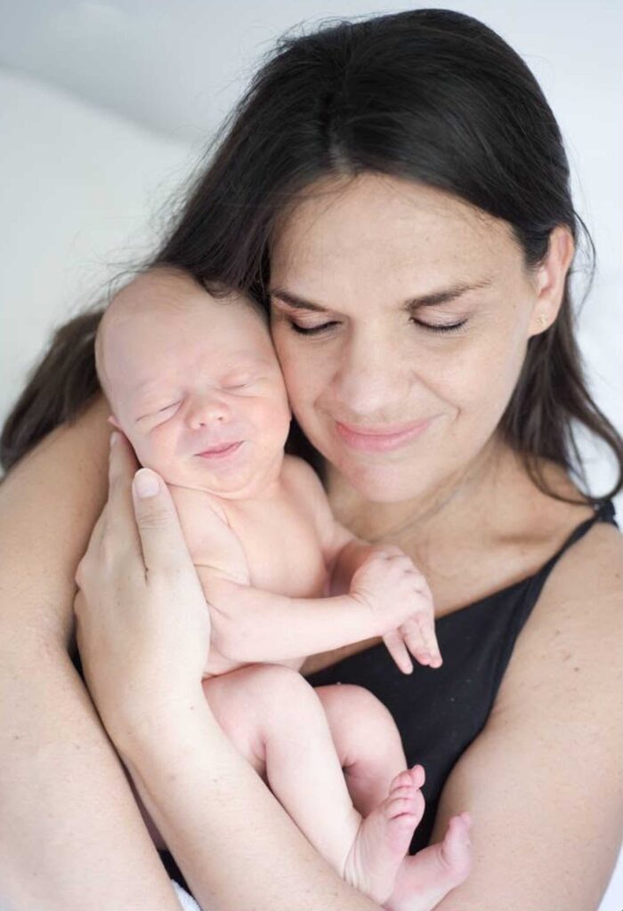 Smiling mother with long dark hair and closed eyes holding her sleeping newborn baby cheek to cheek against her chest, photographed by Adri Krige, newborn photographer in St Francis Bay