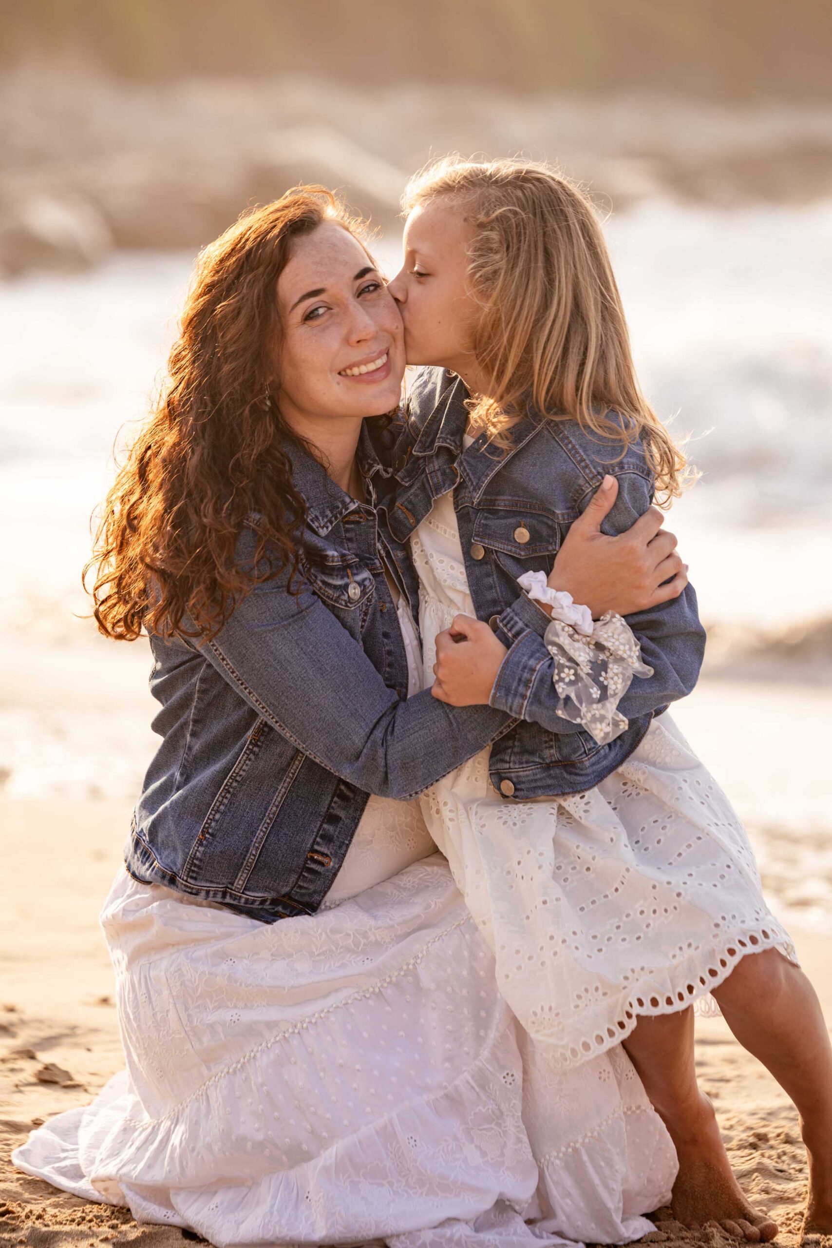 Young girl kissing her smiling mother on the cheek as they hug on the beach at golden hour, both in white dresses and denim jackets, family photography in St Francis Bay by Adri Krige