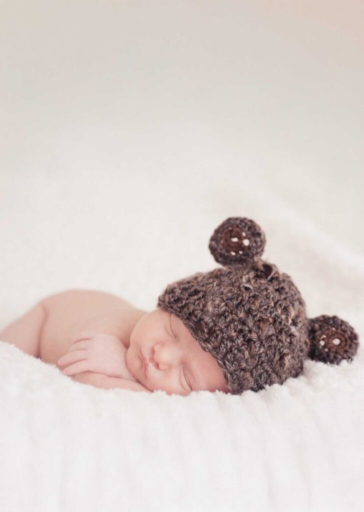 Sleeping newborn baby wearing a brown crochet hat with little bear ears, curled up with tiny hands tucked under the cheek on a soft white blanket, photographed by Adri Krige Photography