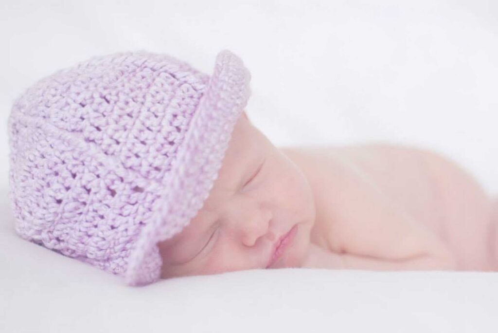 Close-up of a sleeping newborn baby wearing a soft lilac crochet hat, cheek resting on a white blanket in bright, airy light, photographed by Adri Krige Photography