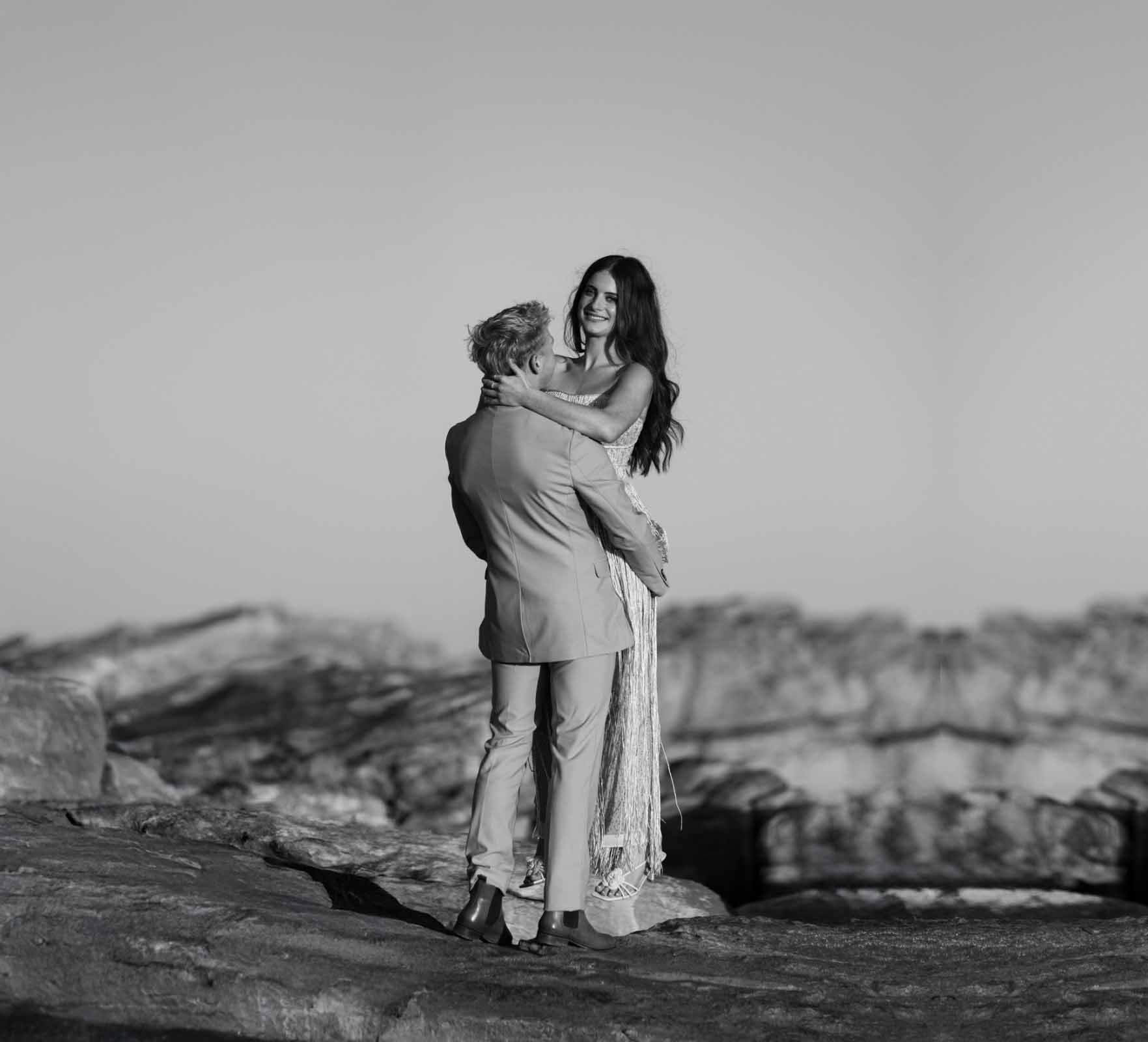 Smiling woman with long dark hair embraced by her fiancé in a suit on coastal rocks, black and white engagement photograph by Adri Krige