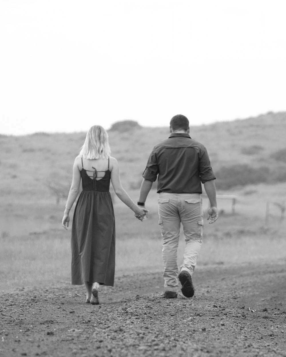 Couple walking hand in hand along a country road, photographed from behind, black and white couples photograph by Adri Krige
