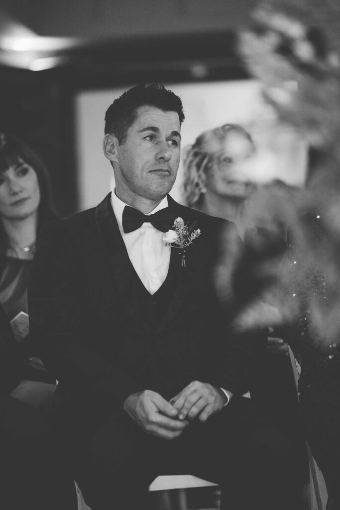 Black and white photo of a man in a bow tie and buttonhole sitting among the guests, watching the wedding ceremony with emotion, at St Francis Links, photographed by Adri Krige, wedding photographer