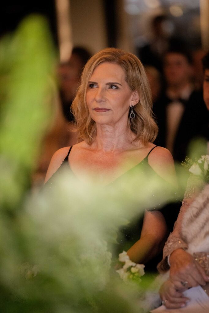 Woman with shoulder-length blonde hair in a black dress watching the wedding ceremony through soft green flowers, at St Francis Links, photographed by Adri Krige, wedding photographer