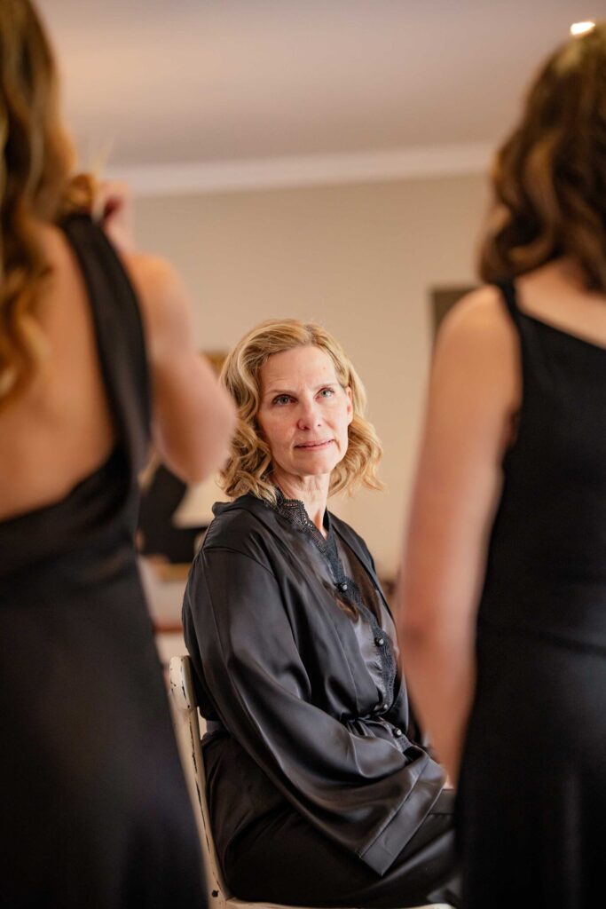 A quiet moment while getting ready Woman with soft blonde curls in a black satin robe sitting on a chair and smiling gently as two women in black dresses get ready in front of her, photographed by Adri Krige, wedding photographer St Francis Bay