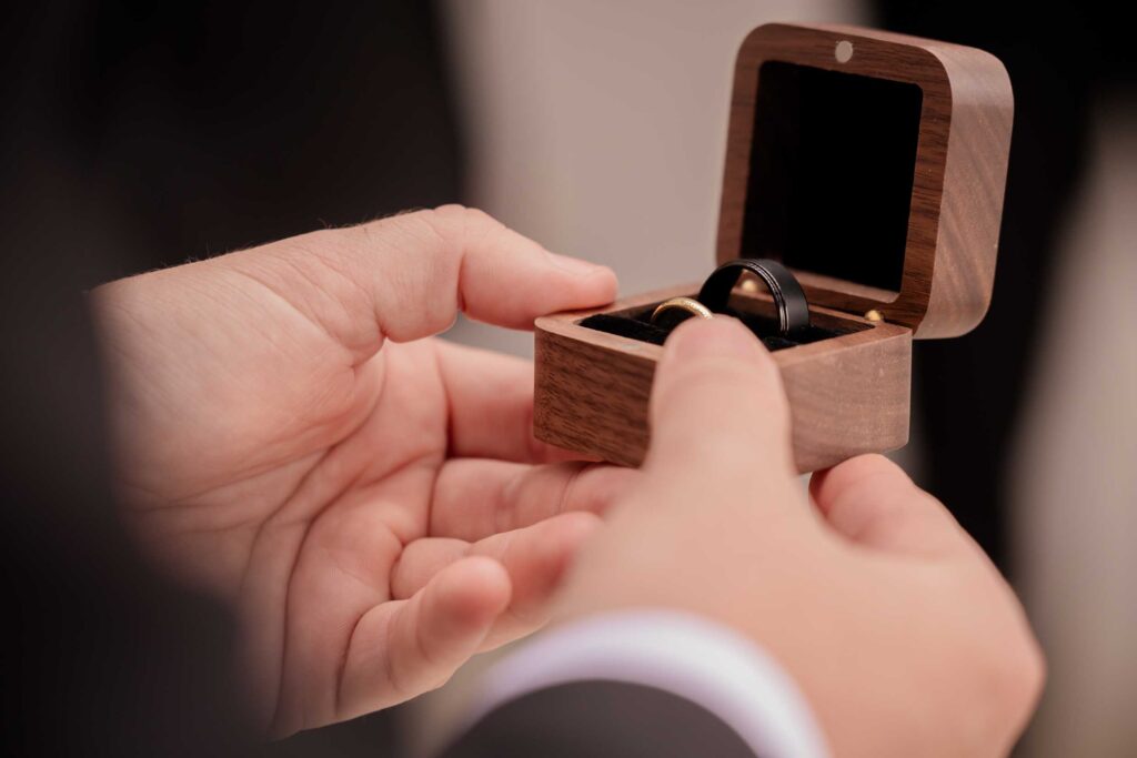 Wedding rings in a wooden box Close-up of hands holding an open wooden ring box with a gold wedding band and a black wedding band inside, photographed by Adri Krige, wedding photographer St Francis Bay
