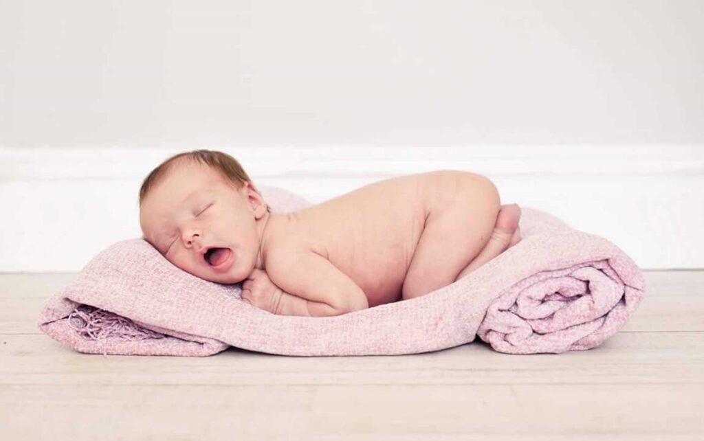 Sleeping newborn baby curled up on a soft pink knitted blanket on a pale wooden floor, mid-yawn with tiny hands tucked under the chin, photographed by Adri Krige Photography